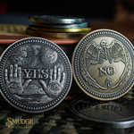 Two metallic challenge coins with engraved designs, one showing 'YES' with hands and suns, and the other showing 'NO' with a winged skeleton, displayed on a dark textured surface with stacked coins in the background. The Smudge logo is visible in the bottom-left corner.