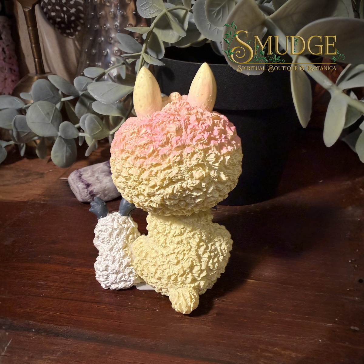 The back of the paco figurine, a yellow lama with a pink top and a small sheep sitting besides him in a botanical background.