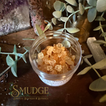 A small clear plastic jar filled with amber-orange crystal bath salts, styled on a dark wooden surface with green leaves, crystals, and a decorative logo for 'SMUDGE Spiritual Boutique & Botanica' at the bottom.