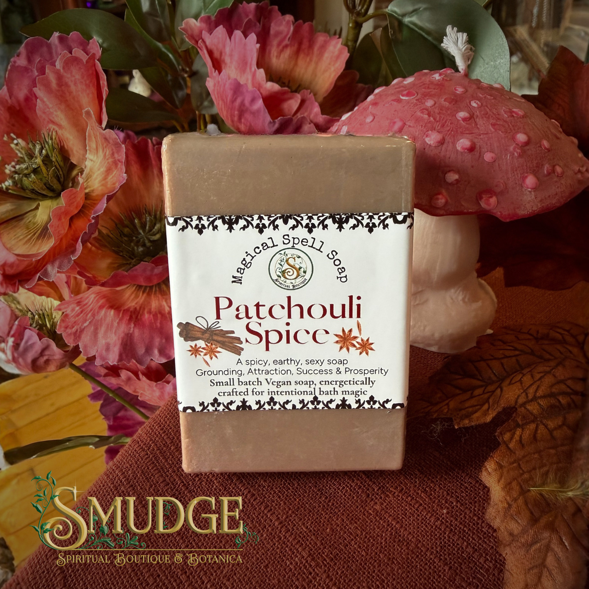 A rectangular bar of 'Patchouli Spice' natural spell soap by Smudge Spiritual Boutique & Botanica, featuring a white label with black and orange text, set against a background of pink flowers and a pink mushroom.
