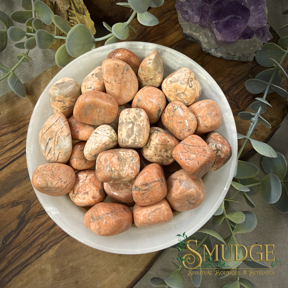 A white ceramic bowl filled with polished peach and beige tumbled stones, styled on a wooden surface with green foliage and crystals in the background. The brand logo 'SMUDGE' is visible in the bottom right corner.