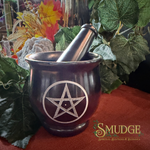 A dark metallic purple/black mortar and pestle set with a silver pentagram symbol, styled with green leaves and crystals on a red cloth surface. The brand logo 'SMUDGE' is visible in the bottom right corner.