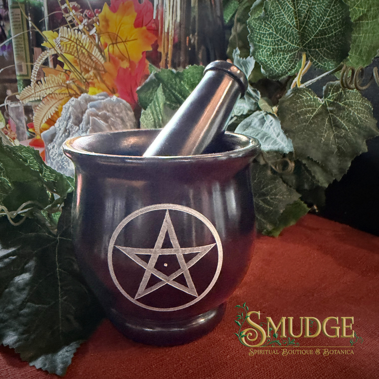 A dark metallic purple/black mortar and pestle set with a silver pentagram symbol, styled with green leaves and crystals on a red cloth surface. The brand logo 'SMUDGE' is visible in the bottom right corner.