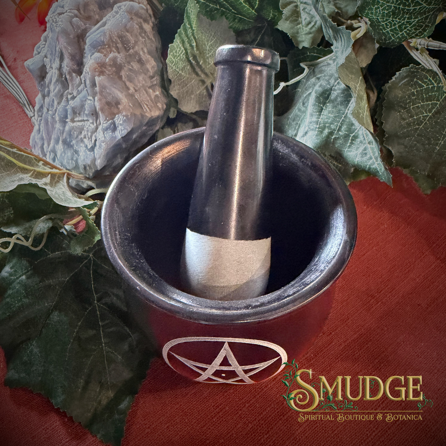 A dark metallic mortar and pestle set with a white pentagram symbol on the outside, placed on a red surface with decorative green leaves and crystals. The brand name 'SMUDGE' is visible in the bottom right corner.
