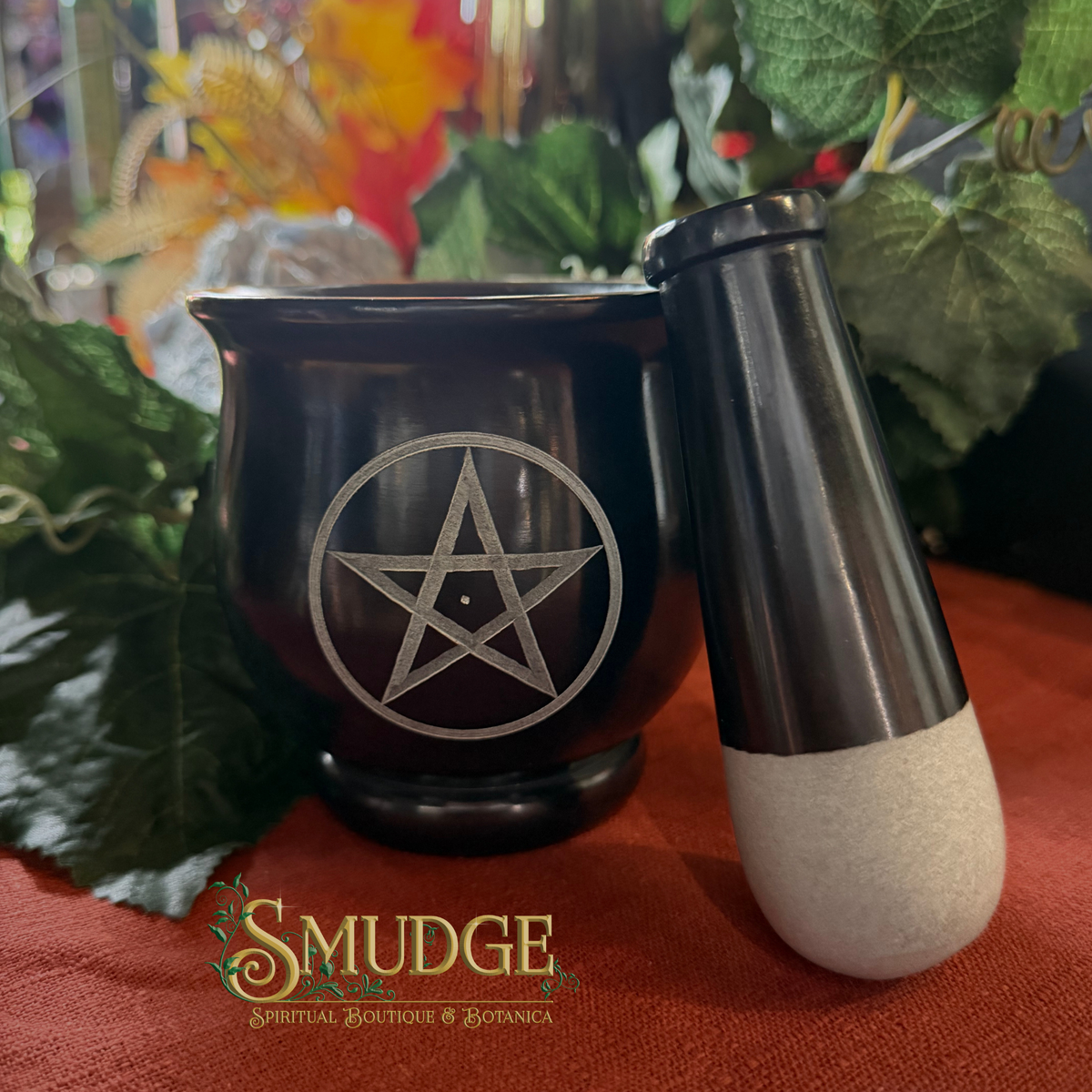 A black ceramic witch's cauldron with a silver pentagram symbol, shown next to a black and white stone pestle on a red cloth with green foliage in the background. The brand logo 'SMUDGE' is visible at the bottom.