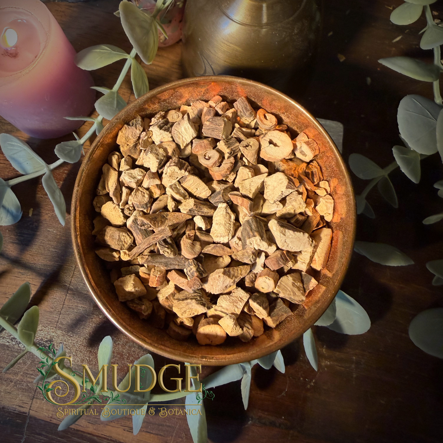 A top-down view of a rustic bronze-colored bowl filled with dried botanical bark pieces, surrounded by eucalyptus leaves on a dark wooden surface. A pink candle and a brass pot are visible in the background, with a 'SMUDGE' brand logo overlay at the bottom left.