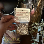 A hand holds a small clear plastic bag of dried peony root herbs with a cream paper header card labeled 'Magical Herbs' and 'Peony Root'. The background is a dark, mystical setting with dried botanicals, a crystal, and a blurred face.