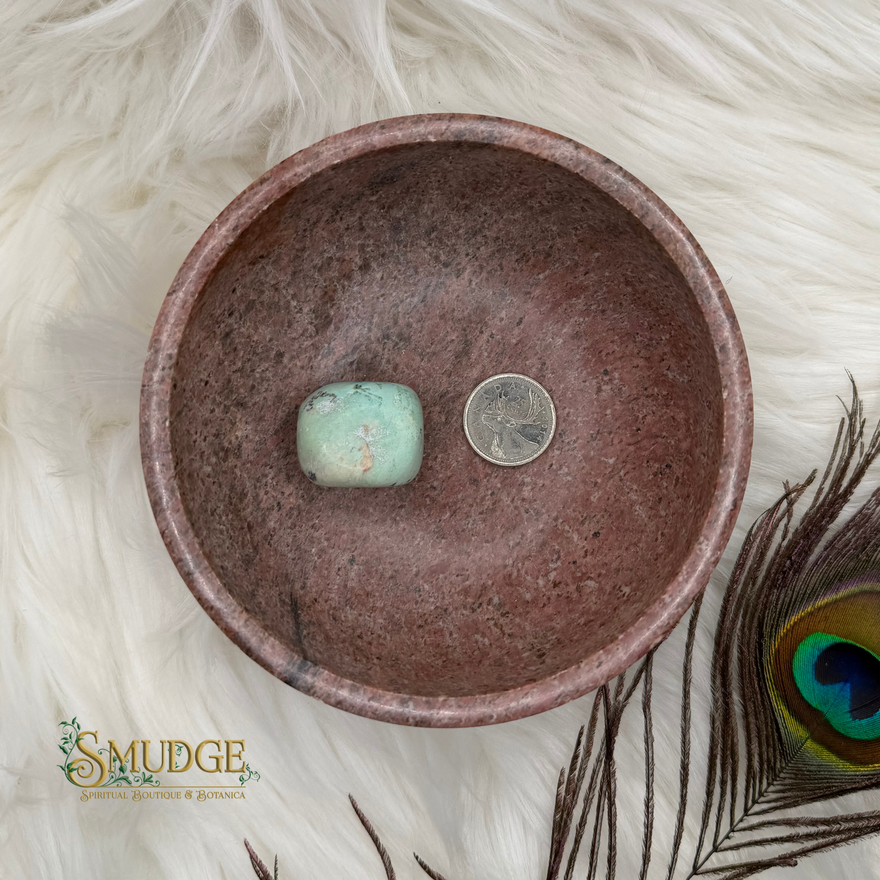 A round, rustic brown ceramic bowl with a speckled finish, shown from a top-down view on a white faux fur surface. Inside the bowl are a light green stone and a silver coin for scale.