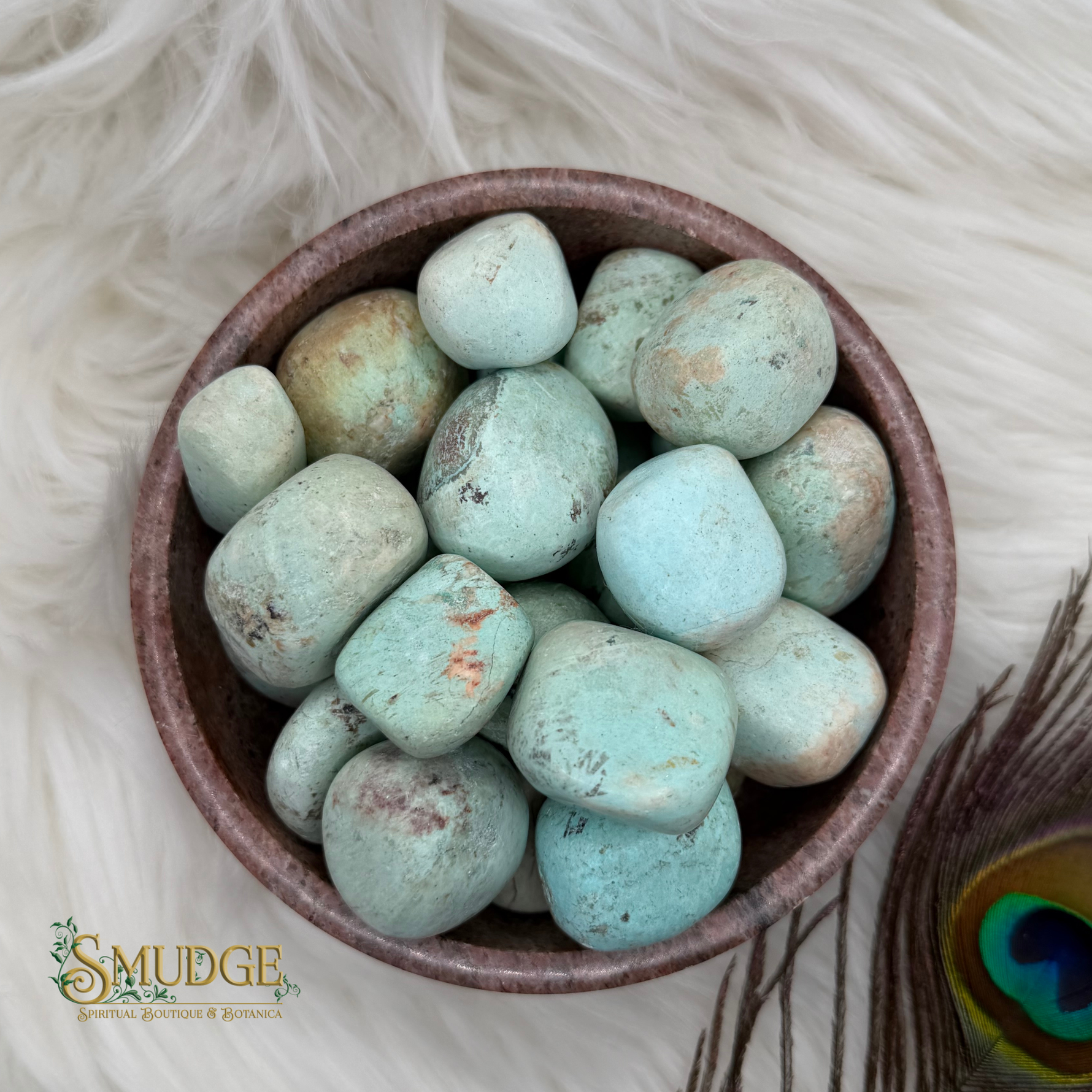 A top-down view of smooth, rounded stones in a brown ceramic bowl, set on a white faux fur background with a peacock feather and the 'SMUDGE Spiritual Goods & Jewelry' logo in the corner.