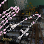 A silver rosary necklace with pink faceted beads and a detailed crucifix pendant, shown draped over a shop counter with shelves of candles in the background. The brand logo 'SMUDGE' is visible in the bottom-left corner.