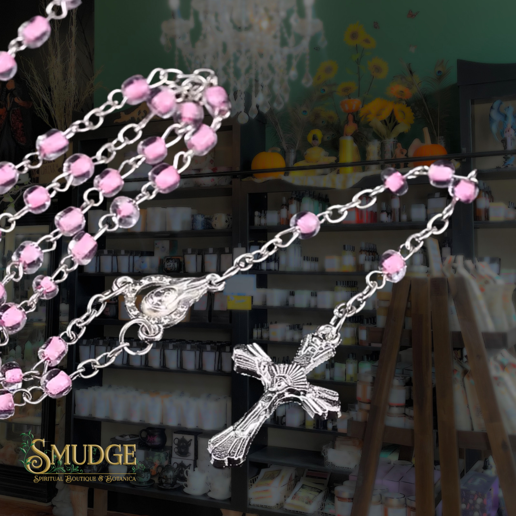 A silver rosary necklace with pink faceted beads and a detailed crucifix pendant, shown draped over a shop counter with shelves of candles in the background. The brand logo 'SMUDGE' is visible in the bottom-left corner.