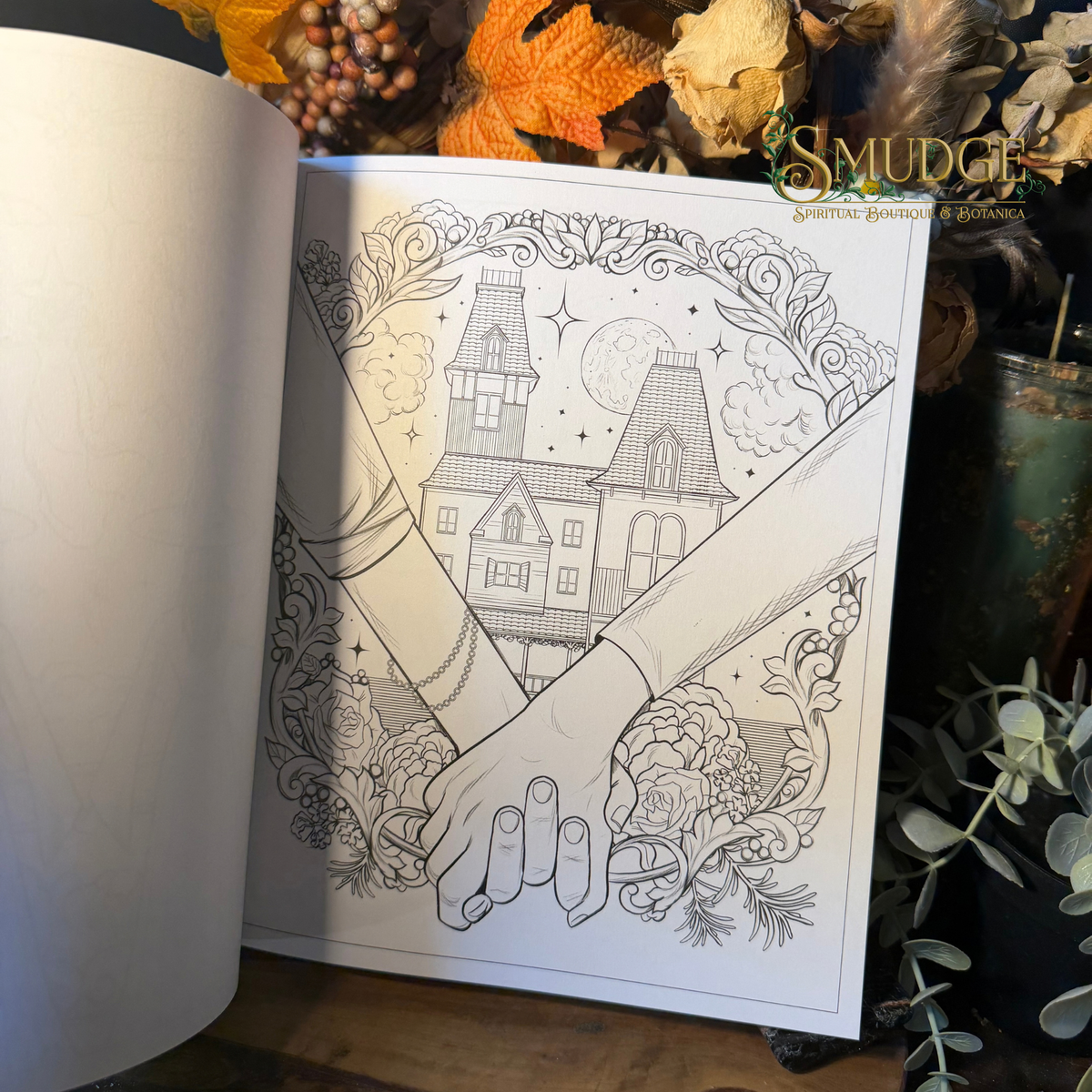 An open art journal or coloring book showing a black-and-white line art illustration of two hands holding, with a haunted house, moon, stars, and ornate floral details. The page is photographed on a tabletop with dried leaves, greenery, and a candle in a cozy styled setting.