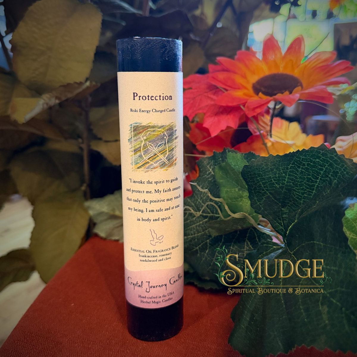 A cylindrical crystal energy candle labeled 'Protection' by Crystal Journey Candles, standing upright on a red surface with artificial green leaves and a red-orange flower in the background. A 'SMUDGE Spiritual Boutique & Botanica' watermark is visible on the right.