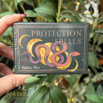 A hand holds a boxed card deck titled "PROTECTION SPELLS" by Judika Illes. The box is dark green with a stylized snake illustration in pink, purple, and gold, and the product is photographed in front of green foliage with warm bokeh lights.