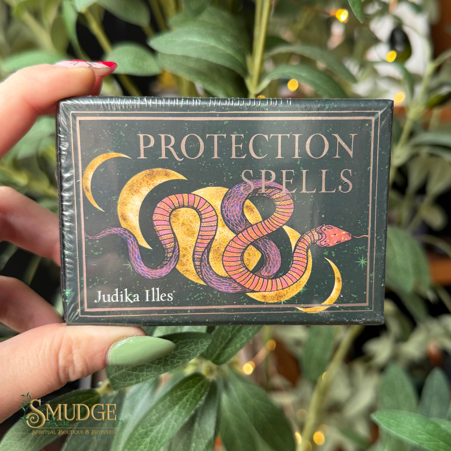 A hand holds a boxed card deck titled "PROTECTION SPELLS" by Judika Illes. The box is dark green with a stylized snake illustration in pink, purple, and gold, and the product is photographed in front of green foliage with warm bokeh lights.