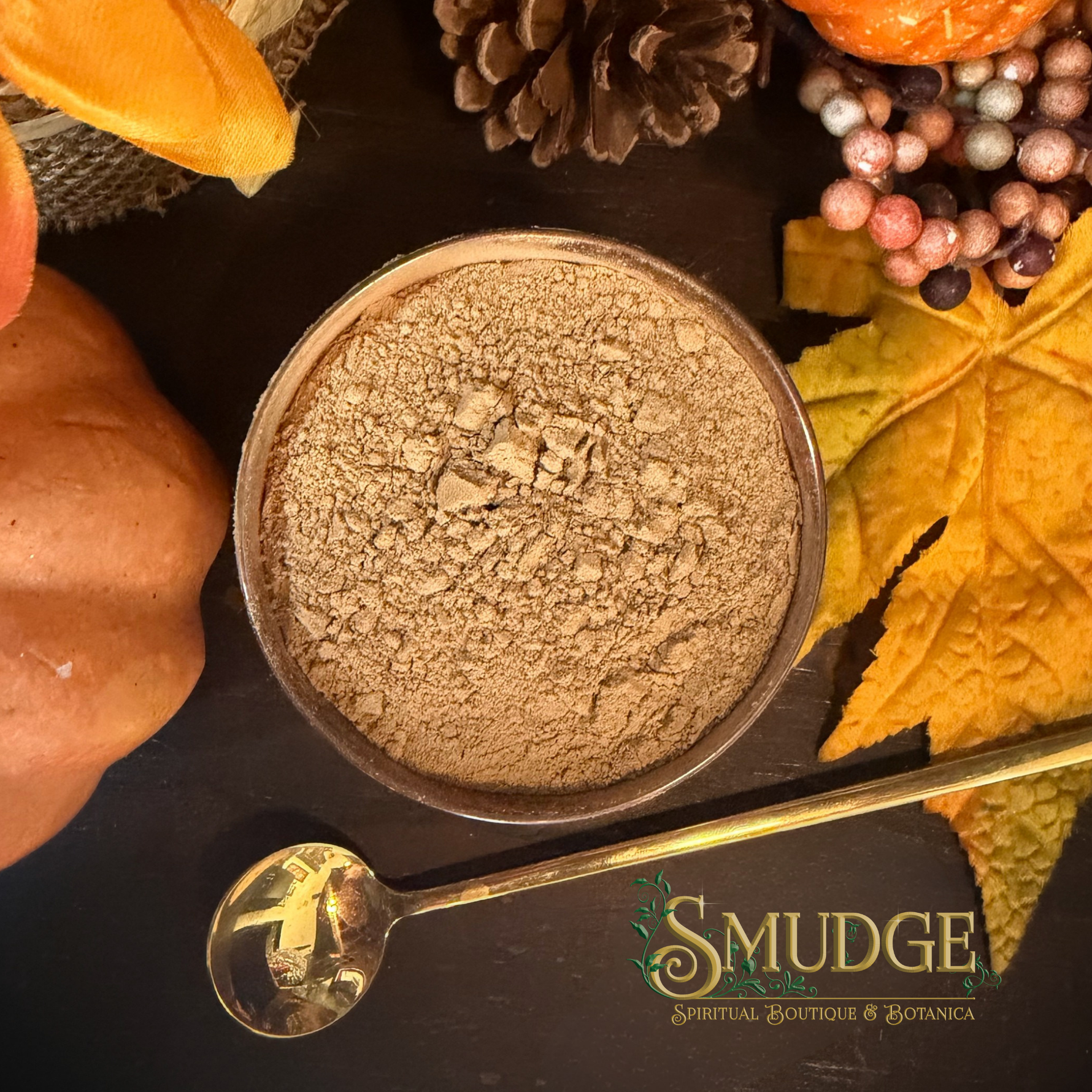 A top-down view of a small metal bowl filled with a light brown powder, surrounded by autumn props including pumpkins, a pinecone, berries, a yellow leaf, and a gold spoon on a dark surface. The brand name 'SMUDGE' is printed on the spoon.