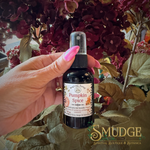 A hand with pink nails holds a small black spray bottle labeled 'Pumpkin Spice' from Sacred Sprigs Botanical Sprigs, set against a background of dark red flowers and green leaves.