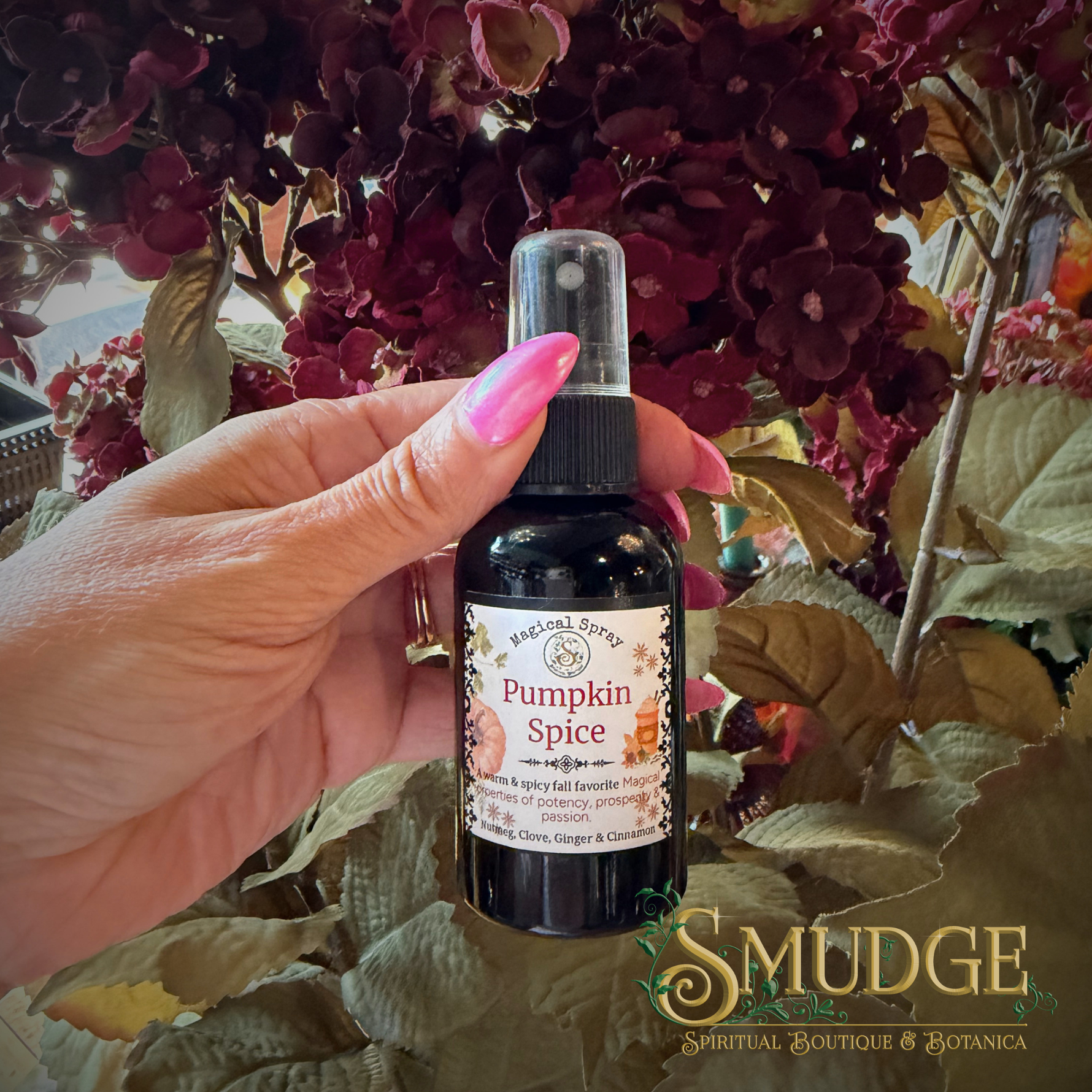 A hand with pink nails holds a small black spray bottle labeled 'Pumpkin Spice' from Sacred Sprigs Botanical Sprigs, set against a background of dark red flowers and green leaves.