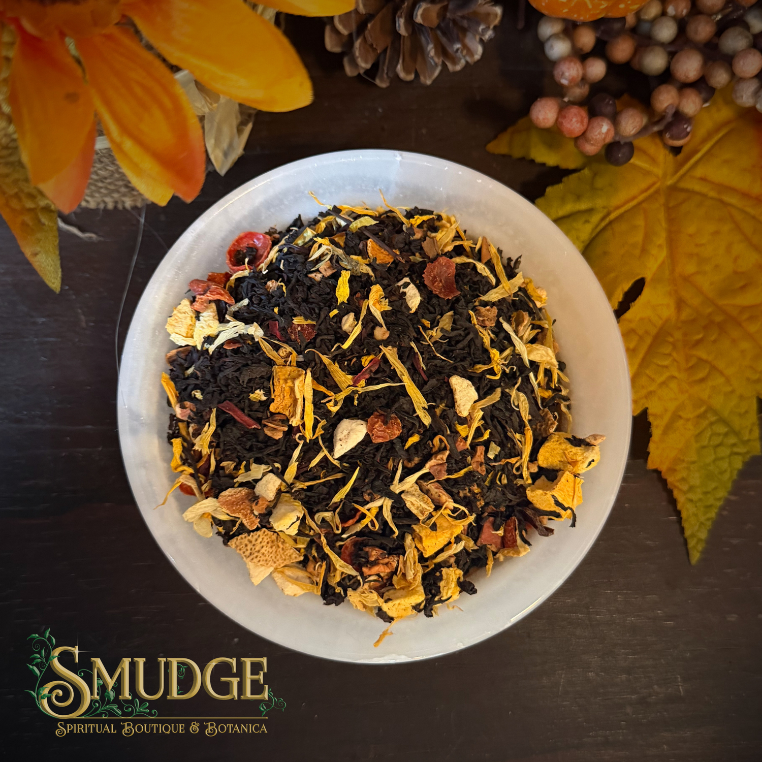 A top-down view of a white bowl filled with a colorful loose-leaf herbal tea blend from Smudge Spiritual Boutique & Botanica, surrounded by autumn leaves, a pinecone, and berries on a dark wooden surface.