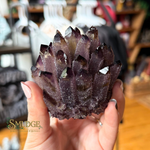 A hand holding a large, rough amethyst crystal cluster with dark purple and black tips, set against a blurred shop background with a 'SMUDGE' watermark in the lower left.