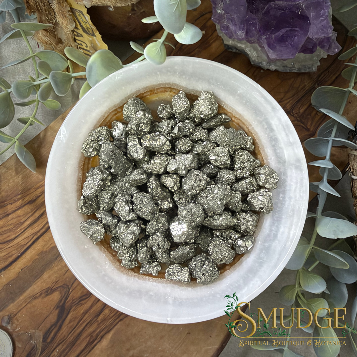 A top-down view of polished metallic pyrite chunks piled in a round white dish, surrounded by decorative greenery and crystals on a wooden surface. The Smudge Spiritual Minerals brand logo is visible in the bottom right corner.