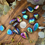A group of polished tumbled iridescent gemstones with a metallic rainbow sheen, arranged on a wooden surface with a silver coin for scale and decorative greenery around them. The stones shift between blue, green, purple, pink, and gold depending on the angle.