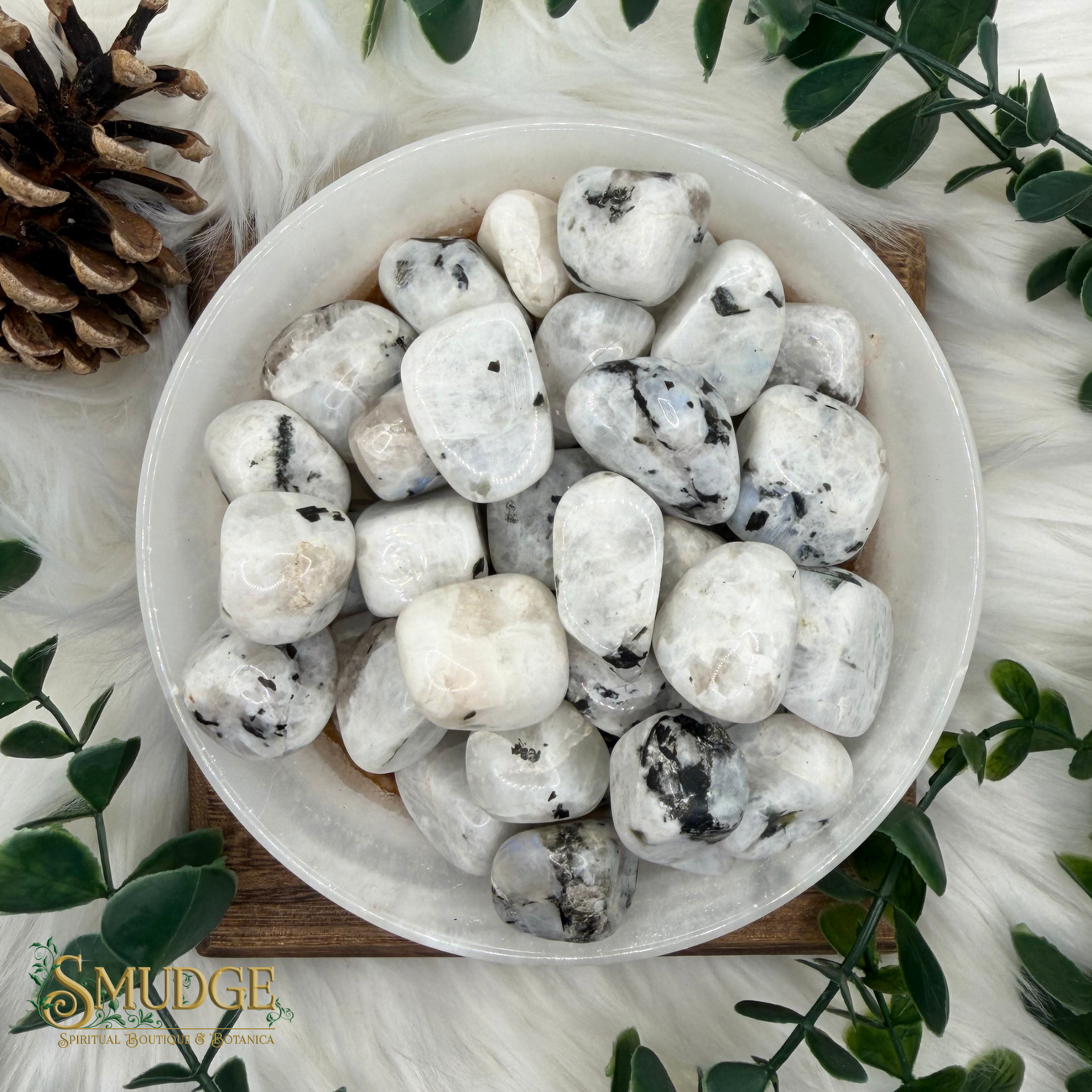 A top-down view of polished white tumbled stones with black and gray veining, arranged in a white bowl on a wooden board, surrounded by green leaves, a pinecone, and white faux fur. The brand logo 'SMUDGE' is visible in the bottom-left corner.