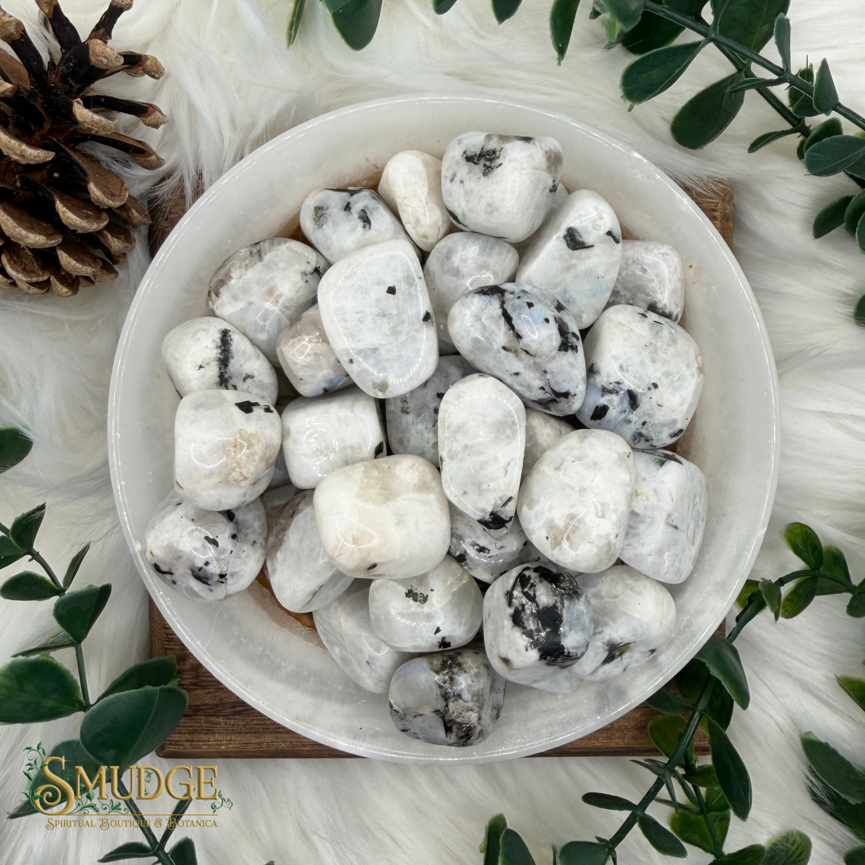 A top-down view of polished white tumbled stones with black and gray veining, arranged in a white bowl on a wooden board, surrounded by green leaves, a pinecone, and white faux fur. The brand logo 'SMUDGE' is visible in the bottom-left corner.