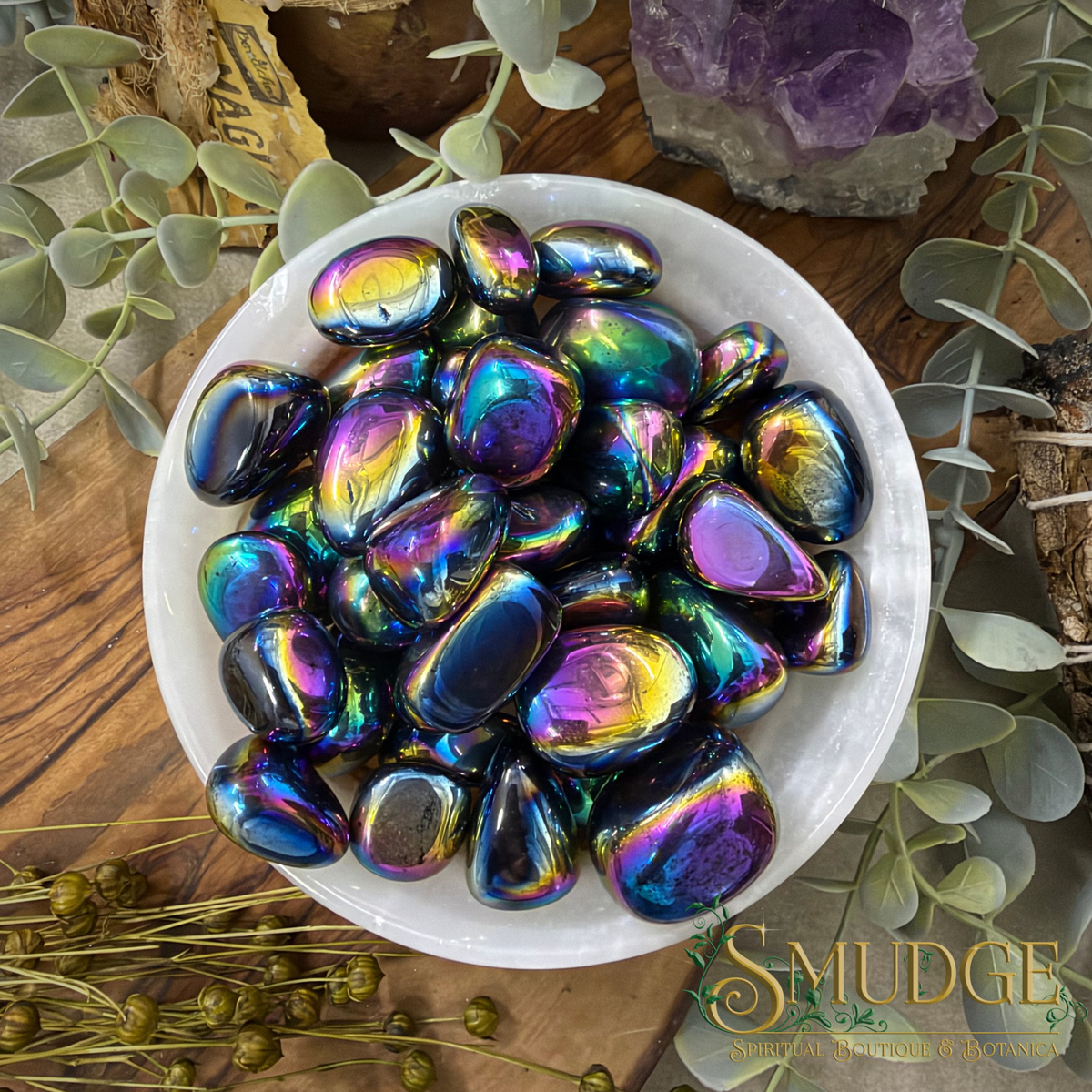 A white bowl filled with smooth, polished tumbled stones that have an iridescent rainbow sheen, set on a wooden surface with decorative greenery and crystals around it. The brand name 'SMUDGE' is visible in the bottom right corner.