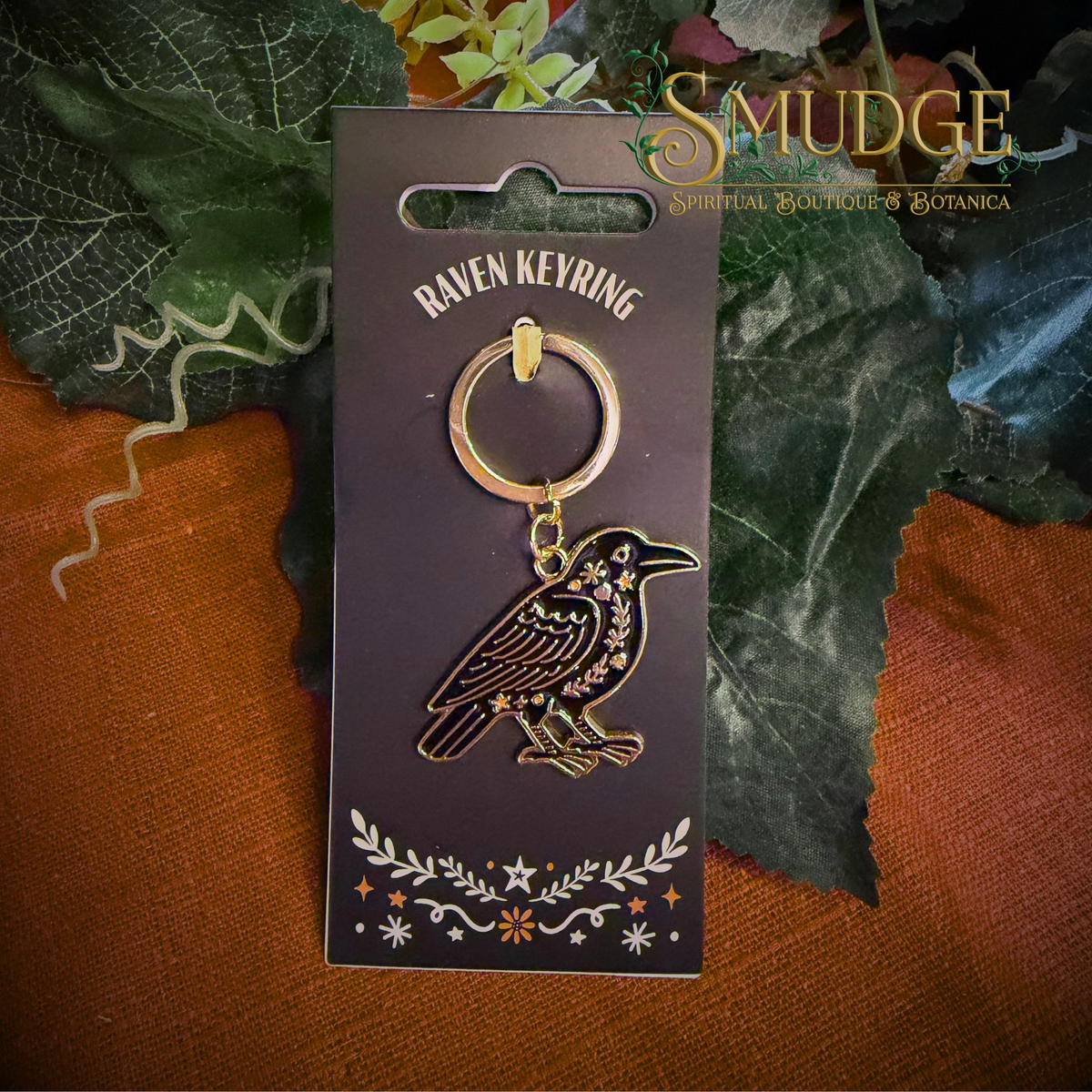 A black and gold raven keyring with floral engravings, displayed on its dark brown backing card. The card is set against a rustic background of orange-brown fabric and green leaves, with the brand name 'SMUDGE' visible in the top right corner.