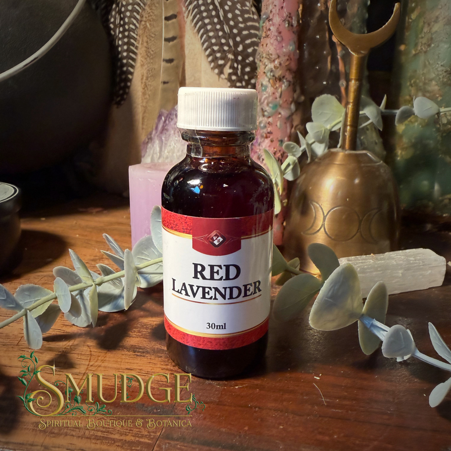 A 30ml amber glass bottle of Red Lavender essential oil or tincture with a white cap and a white label, set on a wooden surface with eucalyptus leaves and mystical props in the background. The brand logo 'Smudge Spiritual Manifest & Botanica' is visible in the bottom-left corner.