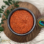 Red spice in a black bowl on a wooden surface with greenery and a peacock feather.