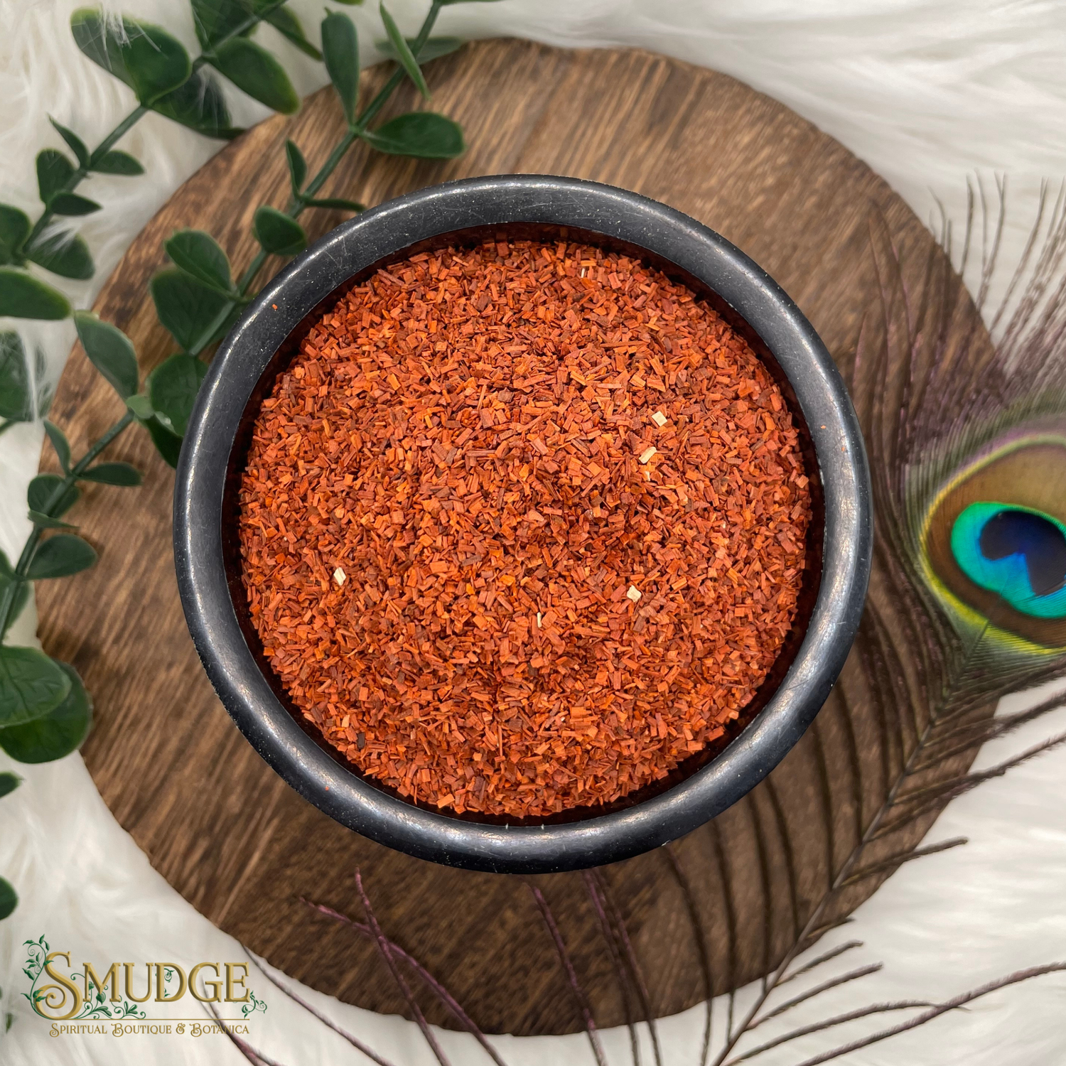 Red spice in a black bowl on a wooden surface with greenery and a peacock feather.