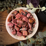 A white ceramic bowl filled with polished pink and reddish rhodonite tumbled stones, photographed from above on a wooden surface with decorative greenery and a purple crystal around it. The words 'SMUDGE SPIRITUAL BOUTIQUE & BOTANICA' appear as a watermark/logo in the lower right corner.