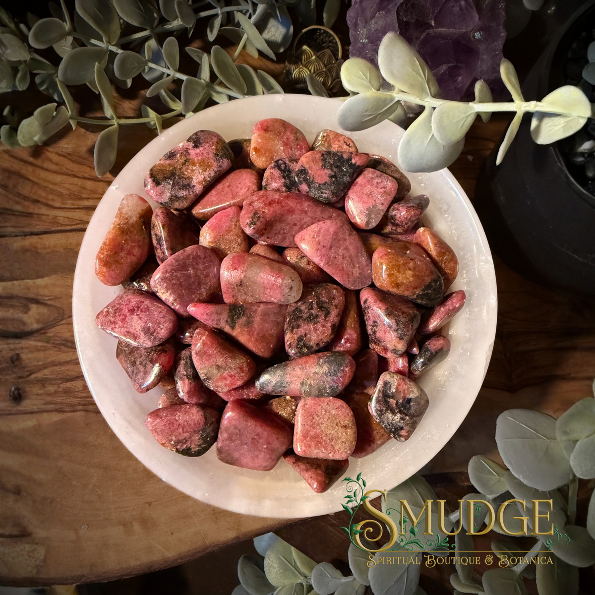 A white ceramic bowl filled with polished pink and reddish rhodonite tumbled stones, photographed from above on a wooden surface with decorative greenery and a purple crystal around it. The words 'SMUDGE SPIRITUAL BOUTIQUE & BOTANICA' appear as a watermark/logo in the lower right corner.
