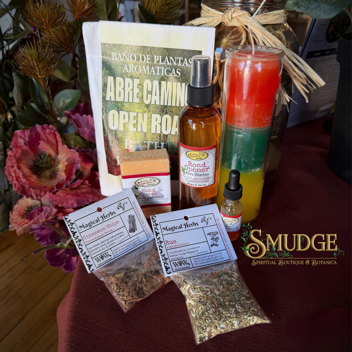 A tabletop display of spiritual and herbal products from Smudge Spiritual Boutique & Botanica, including a 'Rancho de Plantas Aromáticas' book, a red, orange, and green layered candle, a Road to Freedom spray, a small herbal tincture bottle, and two bags of magical herbs on a dark red cloth.
