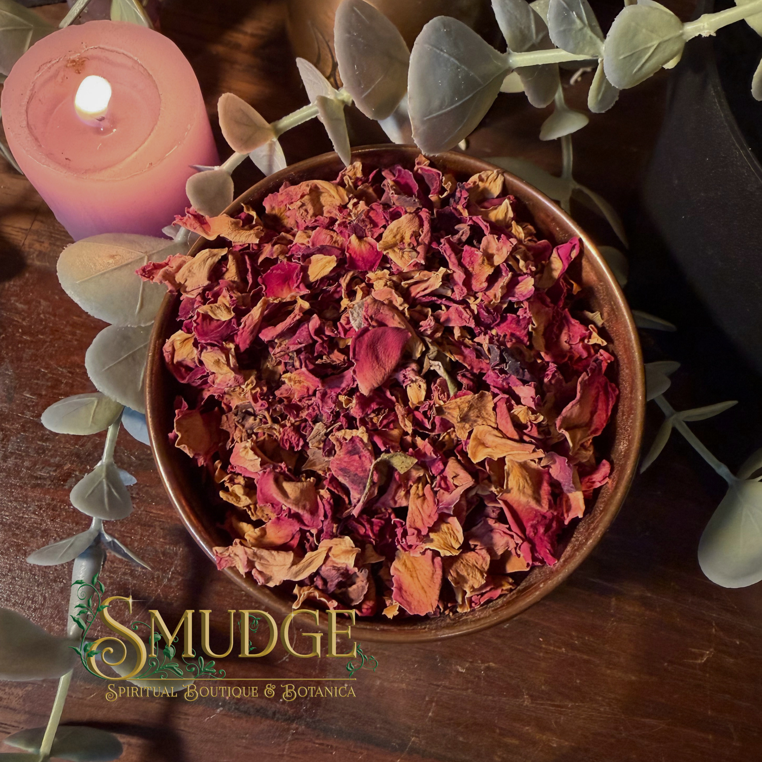A bowl filled with dried rose petals and flower buds, styled on a wooden surface with a pink candle, green eucalyptus leaves, and a visible 'Smudge Spiritual Boutique & Botanica' logo at the bottom.