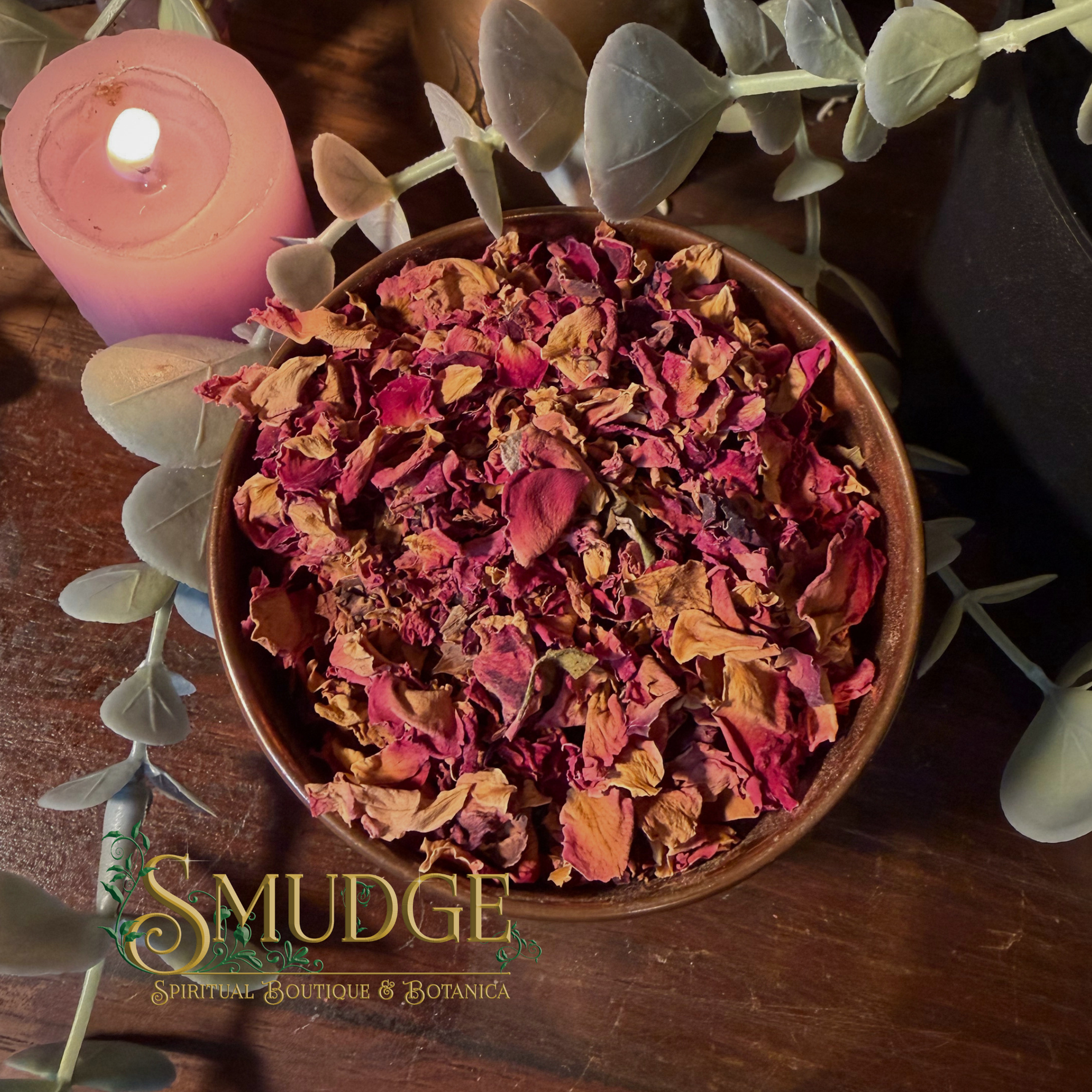 A bowl filled with dried rose petals and flower buds, styled on a wooden surface with a pink candle, green eucalyptus leaves, and a visible 'Smudge Spiritual Boutique & Botanica' logo at the bottom.