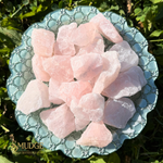 Raw rose quartz crystals in various rough, irregular shapes, displayed on a scalloped light blue ceramic plate set outdoors on green grass. The brand name 'SMUDGE' is visible in the bottom-left corner.