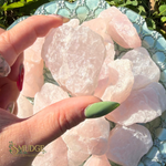 A hand with green and red manicured nails holds a cluster of rough, pale pink rose quartz crystals over a decorative light blue plate outdoors with green leaves in the background. The brand logo 'SMUDGE' is visible in the bottom-left corner.