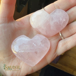 Two faceted rose quartz heart pendants held in a person's hand. The pendants are light pink and translucent, with one showing a natural crystal structure and the other featuring a smooth, polished finish.