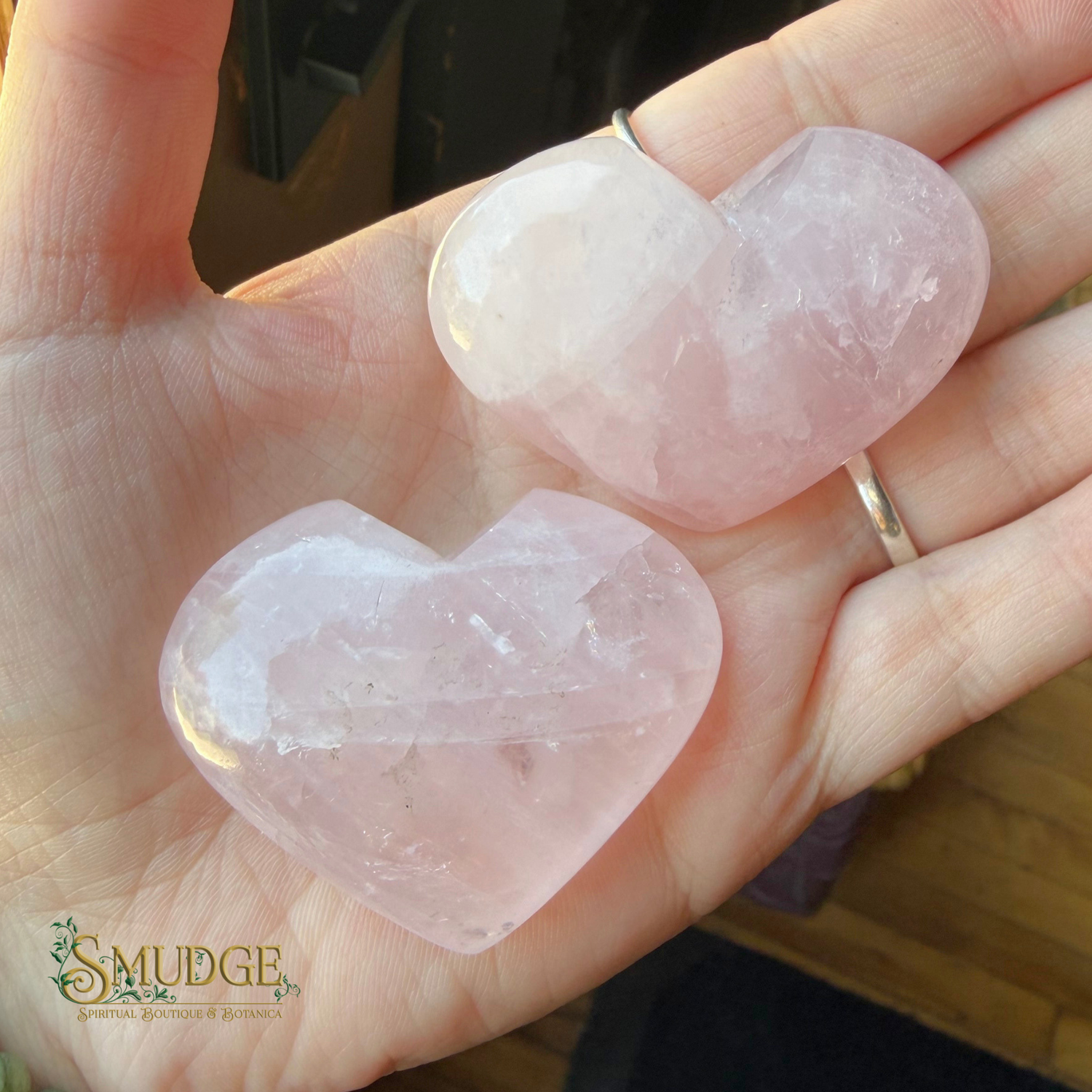 Two faceted rose quartz heart pendants held in a person's hand. The pendants are light pink and translucent, with one showing a natural crystal structure and the other featuring a smooth, polished finish.