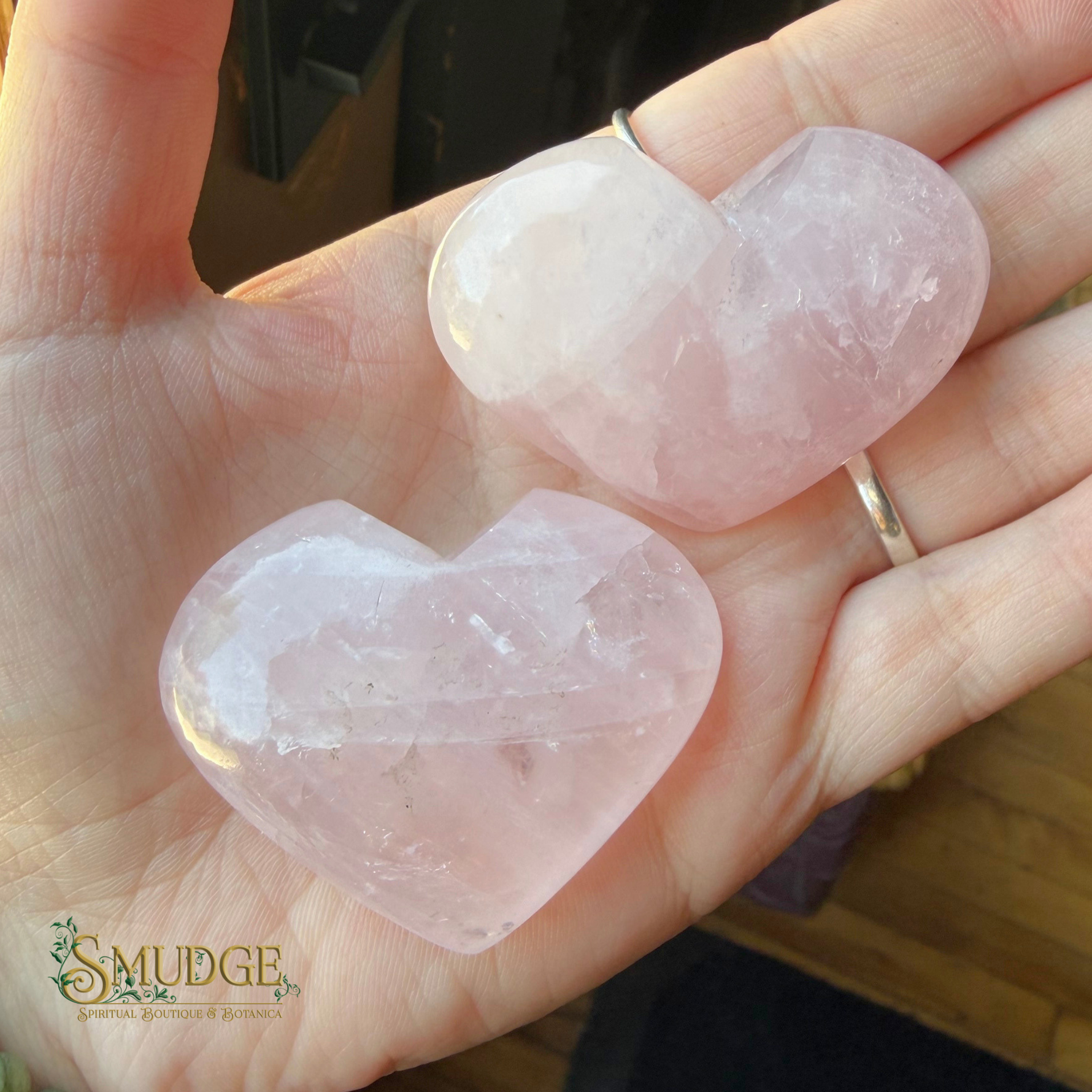 Two faceted rose quartz heart pendants held in a person's hand. The pendants are light pink and translucent, with one showing a natural crystal structure and the other featuring a smooth, polished finish.