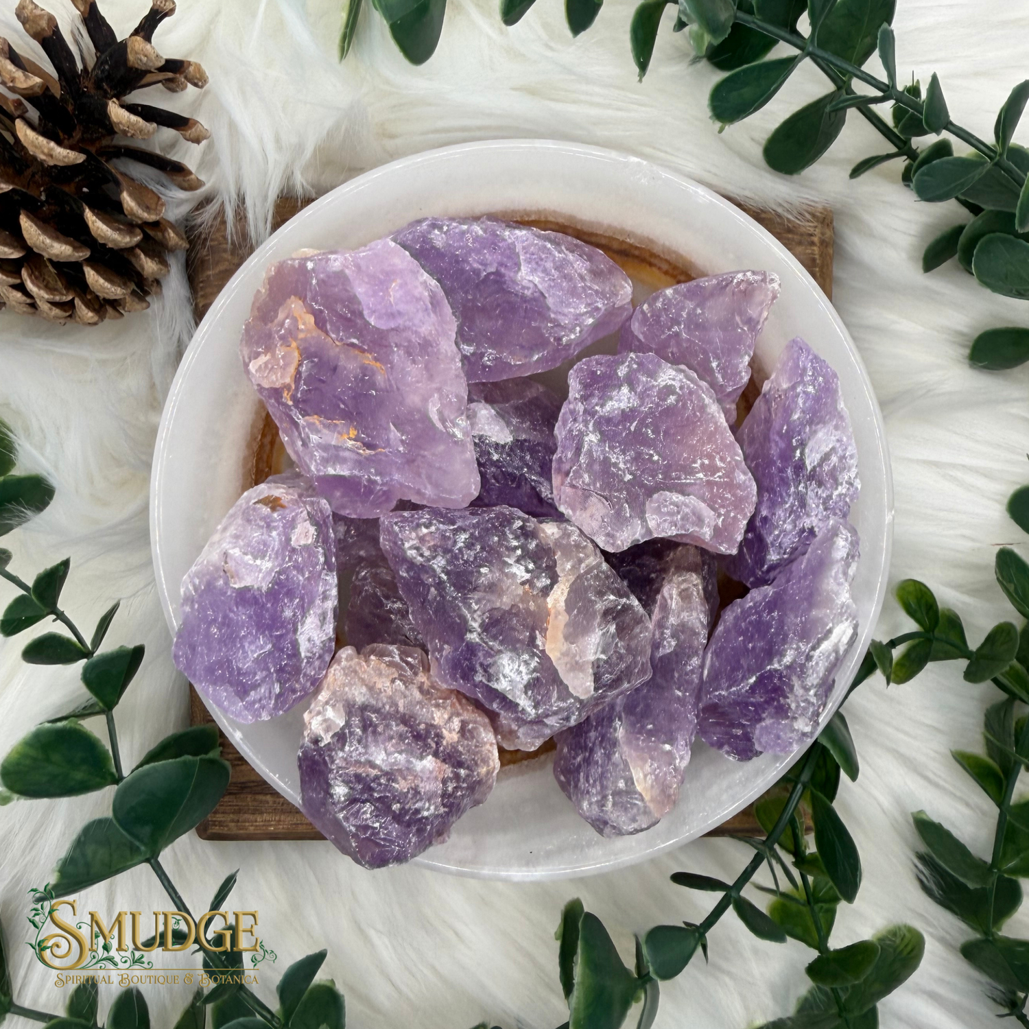 Raw amethyst crystal chunks in a white ceramic bowl, styled with green leaves, a pine cone, and a white faux fur background. The Smudge brand logo is visible in the bottom-left corner.