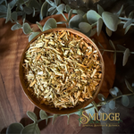 A top-down view of a wooden bowl filled with dried, chopped rue surrounded by green eucalyptus leaves on a dark wooden surface. The brand name 'SMUDGE' is visible in the bottom right corner.