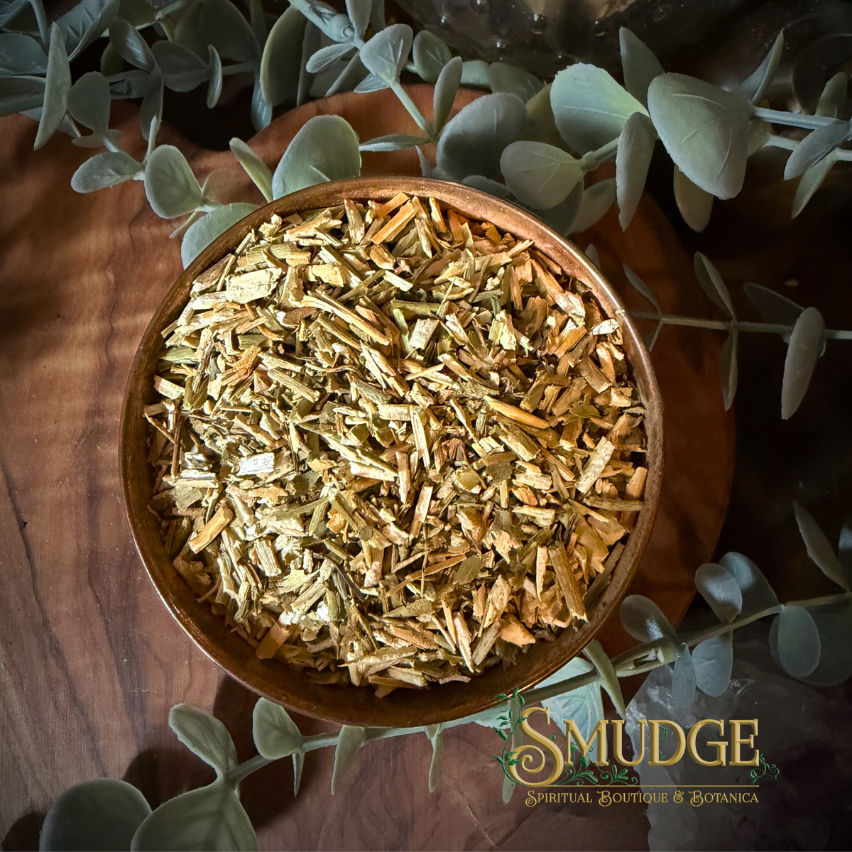 A top-down view of a wooden bowl filled with dried, chopped rue surrounded by green eucalyptus leaves on a dark wooden surface. The brand name 'SMUDGE' is visible in the bottom right corner.