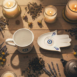 White mug with blue eye design and Hamsa hand-shaped object on a white background