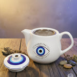 White teapot with eye design on a gray surface, surrounded by a bowl of oranges and a patterned cup.
