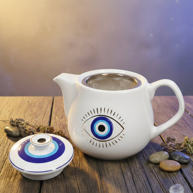 White teapot with eye design on a gray surface, surrounded by a bowl of oranges and a patterned cup.