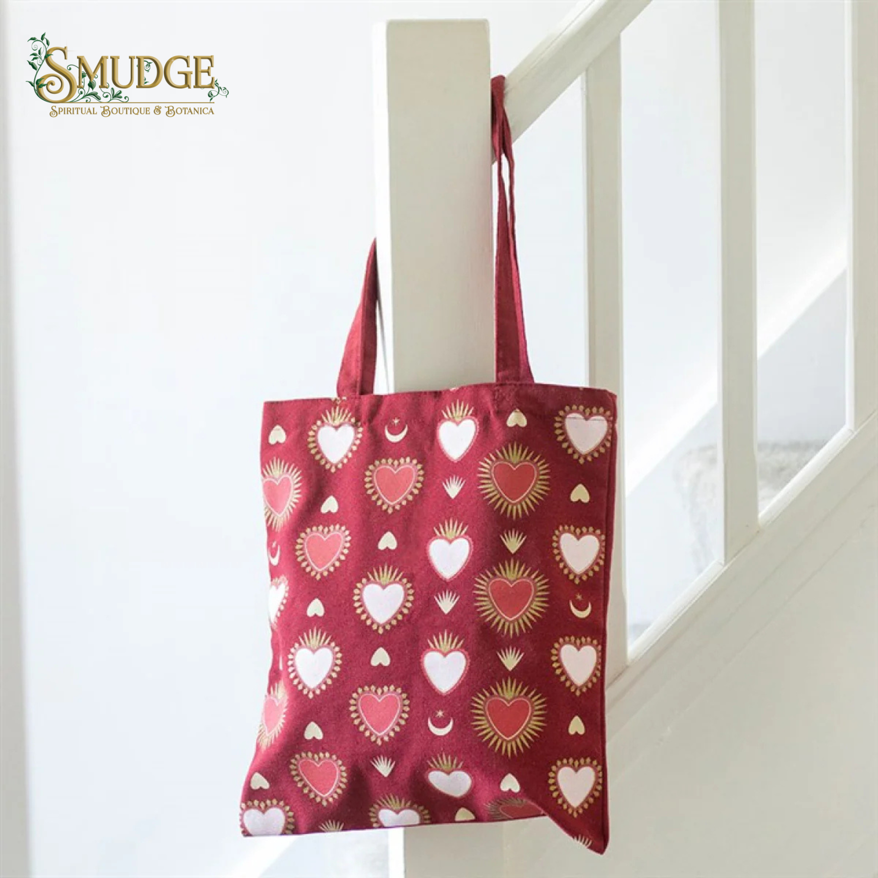 A red canvas tote bag with a repeating heart pattern in pink, red, and white, hanging from a white wooden staircase railing. The Smudge Spiritual Boutique & Botanica logo is visible in the top left corner.